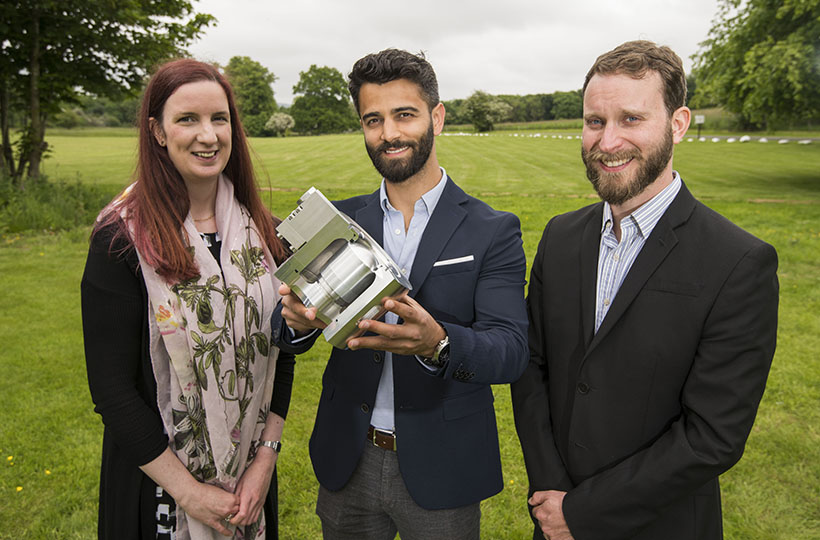 Three people posing for the camera standing side by side in a green field wearing business clothes. Person in the middle is holding a metallic valve.