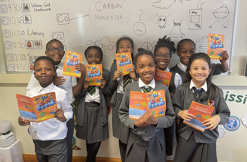 8 school children in grey uniforms stand in front of a white board. They are holding bonanza leaflets and smiling. 