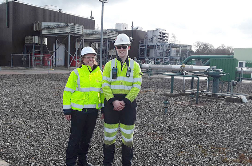 A man and woman in SGN PPE on a pebbled site with multiple pipes