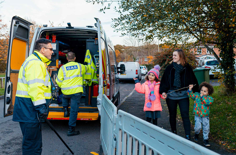 A happy family walking past two SGN engineers working at their van in a street. 