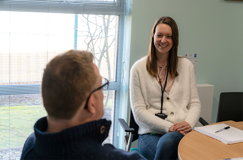 A smiling woman in an office wearing a lanyard speaking to a man