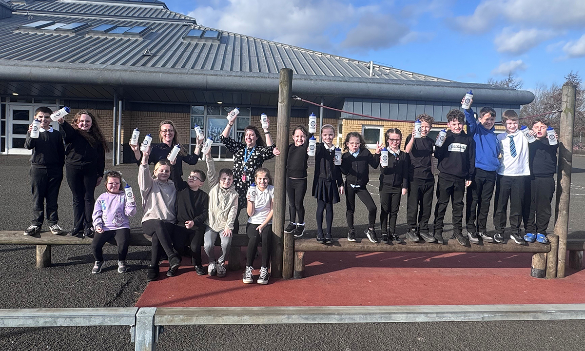 A group of school children outside a school holding up plastic water bottles