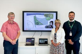 An image with three people posing for camera standing near a monitor