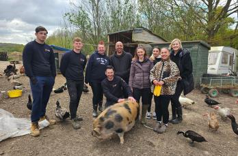 Nine people in coats and tshirts smiling in a muddy location with a pig